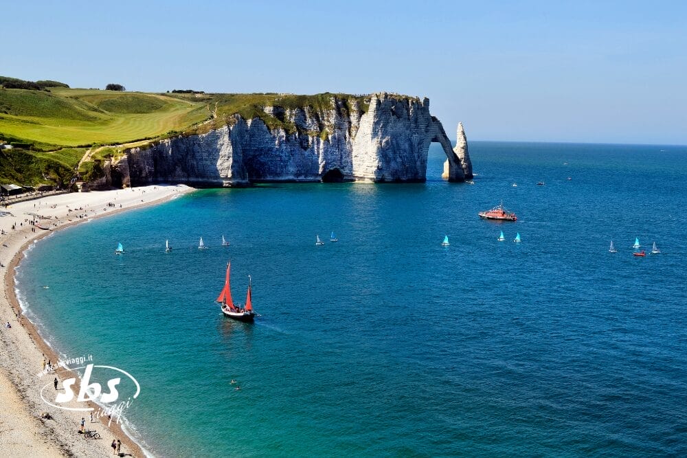 Una costa panoramica con bianche scogliere di gesso, un arco di pietra naturale, verdi colline erbose, una spiaggia di ciottoli, acque turchesi punteggiate di barche a vela e una barca a vele rosse, come una splendida bozza automatica dell'arte della natura.