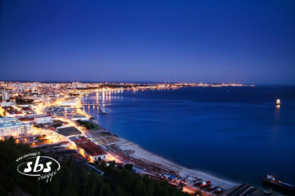 Una città costiera al tramonto nel Sud del Portogallo, con edifici illuminati lungo la costa, mare calmo e blu e cielo che si sta oscurando. È visibile un porto con banchine e barche e il logo "sbs viaggi" in basso a sinistra.