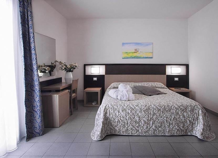 Una moderna camera d'albergo con letto matrimoniale, biancheria da letto a fantasia, due comodini con lampade, una scrivania con specchio e sedia, vasi con fiori e una finestra con tende velate crea un ambiente rilassante Bozza automatica. Un asciugamano è piegato sul letto.