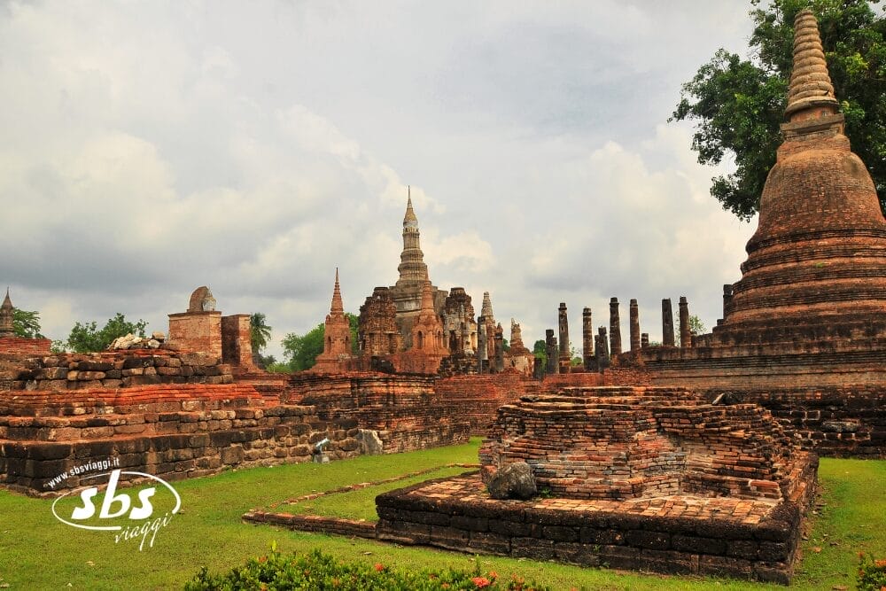 Antiche rovine in mattoni e grandi stupa nel sito di un tempio storico, con erba verde e cielo nuvoloso: una scena suggestiva spesso visitata durante un Tour Thailandia da Nord a Phuket. Filigrana "sbs viaggi" in basso a sinistra.
