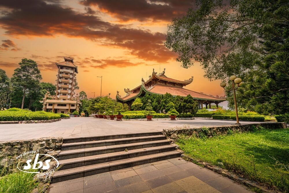 Un tempio tradizionale vietnamita con tetti curvi e una vegetazione lussureggiante, sullo sfondo di un drammatico cielo arancione al tramonto; un alto edificio moderno e una Bozza automatica si trovano nelle vicinanze.