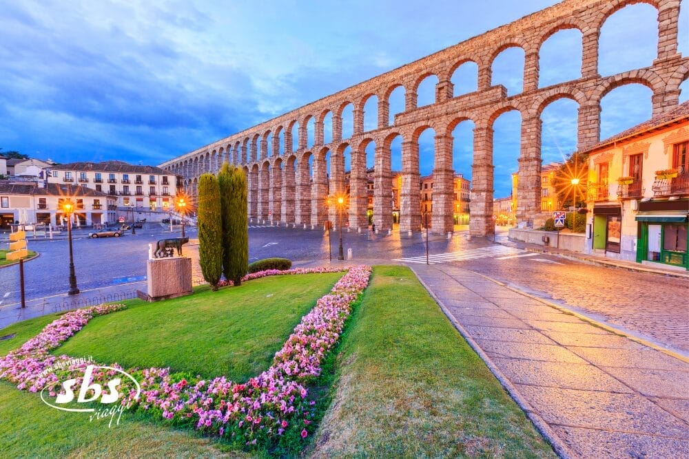 Un ampio acquedotto in pietra con alti archi attraversa una piazza di Segovia, in Castiglia, al tramonto. Aiuole di fiori, una statua e vecchi edifici incorniciano la scena sotto un cielo nuvoloso, con i caldi lampioni che illuminano questo incantevole luogo da minitour di Pasqua.