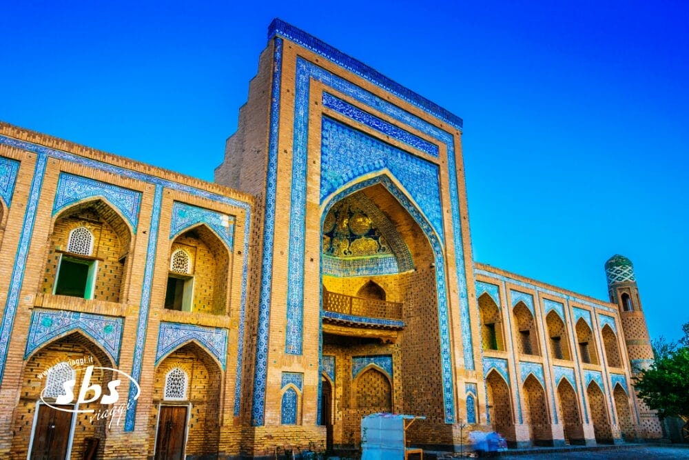 Una grande moschea decorata con intricate piastrelle blu e portali ad arco si staglia contro un cielo azzurro e limpido, come un capolavoro di Bozza automatico. Un alto minareto si erge sulla destra ed eleganti dettagli in mattoni ornano la facciata.