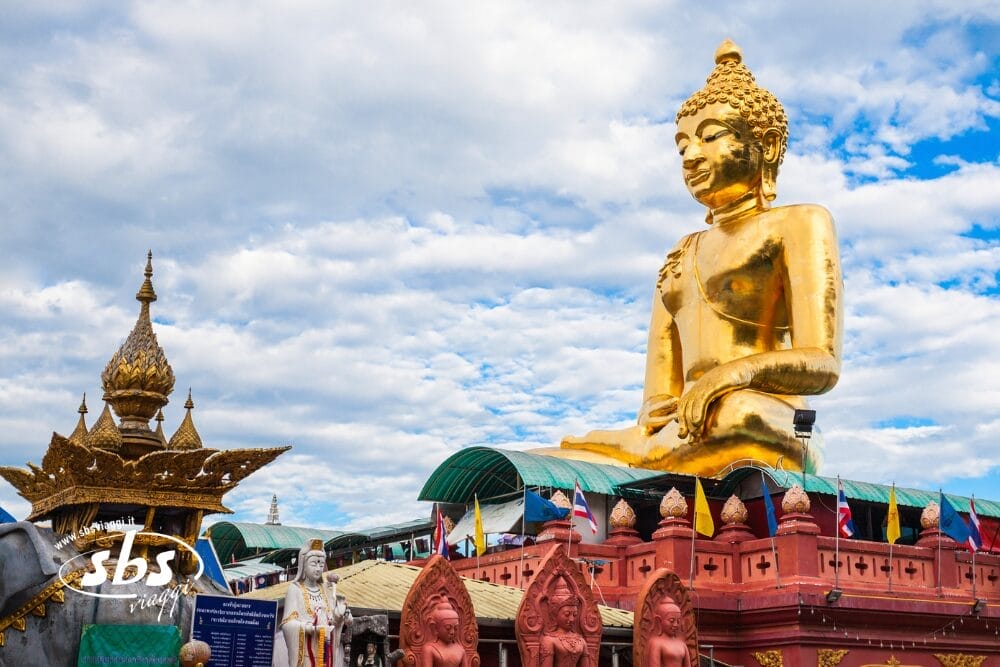 Una grande statua dorata di Buddha si trova all'aperto in un cielo azzurro parzialmente nuvoloso, circondata da strutture ornamentali, bandiere e persone in un complesso di templi, esemplificando la serenità di Bozza automatica.