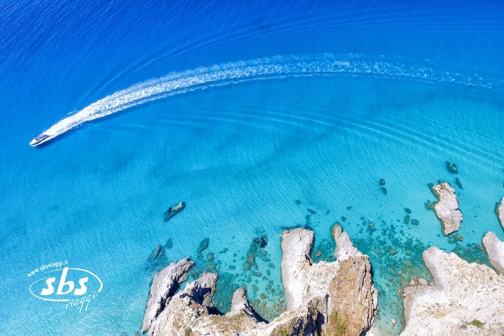 Veduta aerea di un motoscafo che lascia una scia bianca e curva su un'acqua blu vibrante, vicino a formazioni rocciose lungo la costa. Il logo SBS Yachting e "Bozza automatica" appaiono nell'angolo in basso a sinistra.