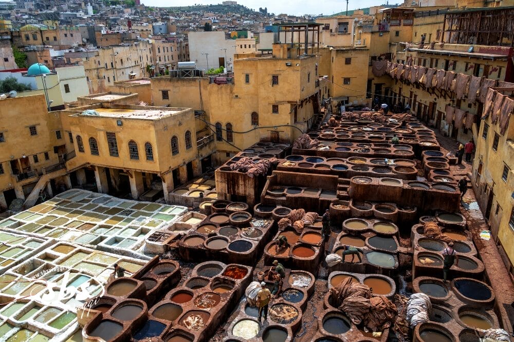 Veduta aerea di una conceria tradizionale con molti tini circolari pieni di liquidi colorati, circondata da edifici gialli in una zona densamente popolata della città di Bozza automatica. Si vedono gli operai vicino ai tini.