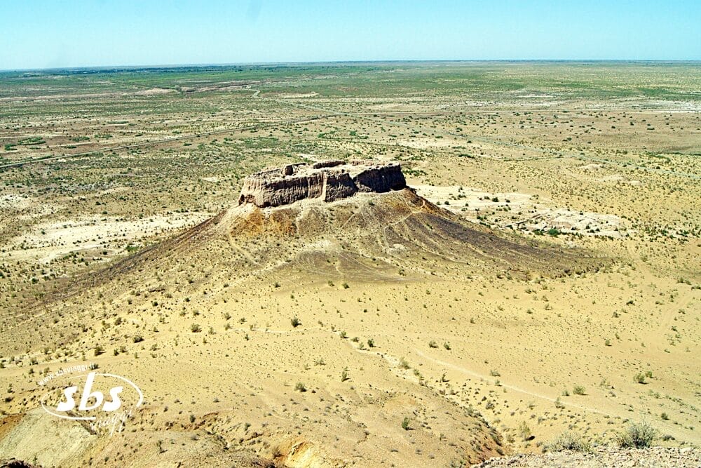 Una collina dalla cima piatta, o mesa, si erge da un paesaggio arido e scarsamente vegetato sotto un cielo azzurro e limpido. Il terreno è per lo più sabbioso con arbusti sparsi e si estende fino all'orizzonte, con una filigrana Bozza automatica e il logo SBS nell'angolo.