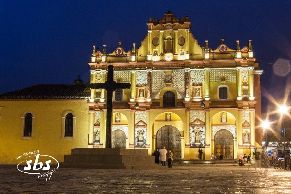 La facciata gialla illuminata della Cattedrale di San Cristóbal de las Casas in Chiapas di notte, con una grande croce di pietra davanti e alcune persone in piedi nella piazza.