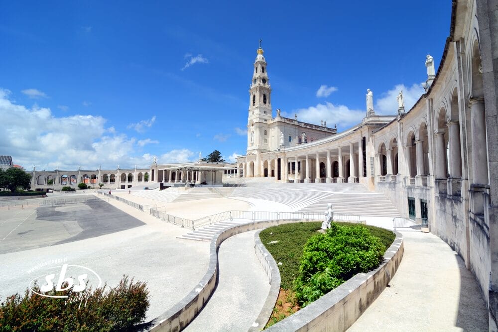 Ampia veduta del Santuario di Fátima in Portogallo, con la sua grande basilica bianca, il colonnato ad archi, il campanile, le scale, il cortile e il cielo azzurro. In primo piano una statua e degli arbusti verdi ripresi come parte di una Bozza automatica.