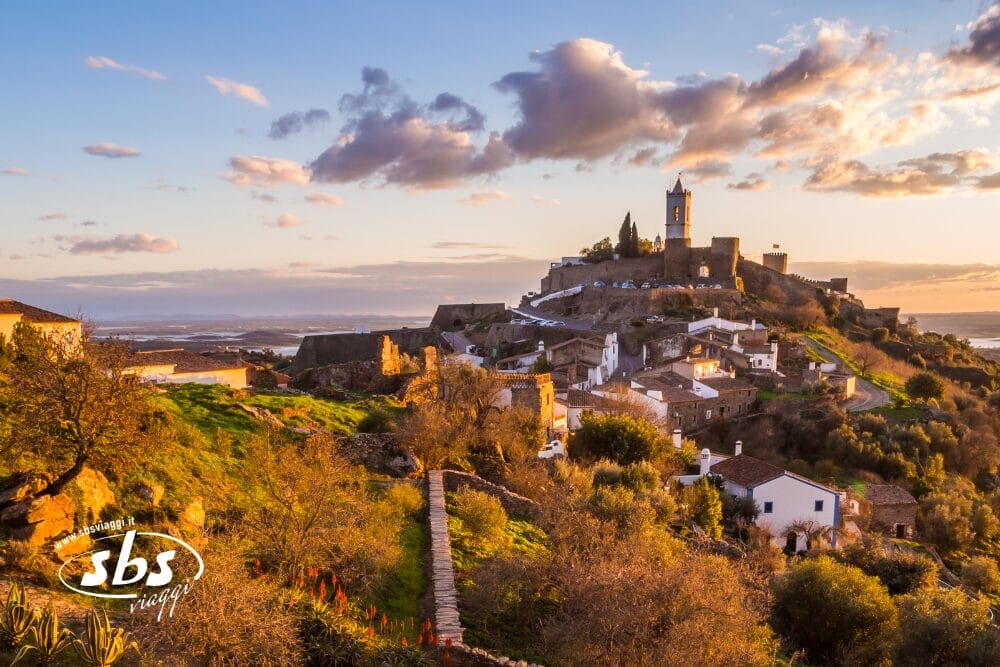 Un villaggio collinare nel Sud del Portogallo, con case bianche e un campanile prominente, è circondato da muri di pietra e verde lussureggiante, il tutto immerso nella luce dorata del sole sotto un cielo parzialmente nuvoloso.