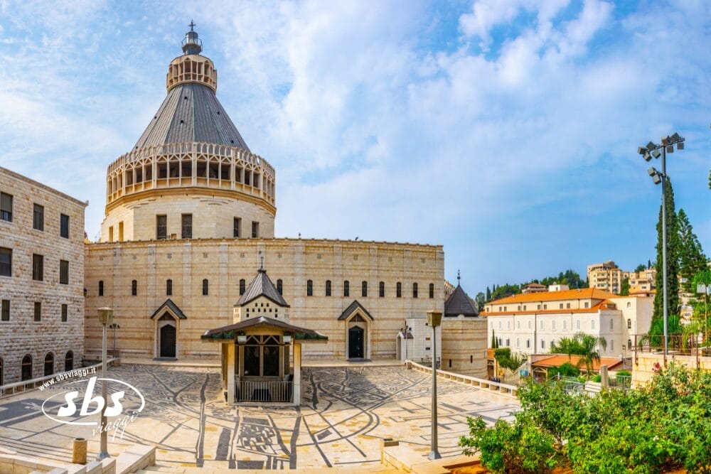 L'immagine mostra la Basilica dell'Annunciazione di Nazareth, in Israele, con la sua grande cupola, la facciata in pietra chiara, il cortile geometrico e gli edifici circostanti sotto un cielo parzialmente nuvoloso: uno spettacolo impressionante, degno di qualsiasi Bozza automatico.