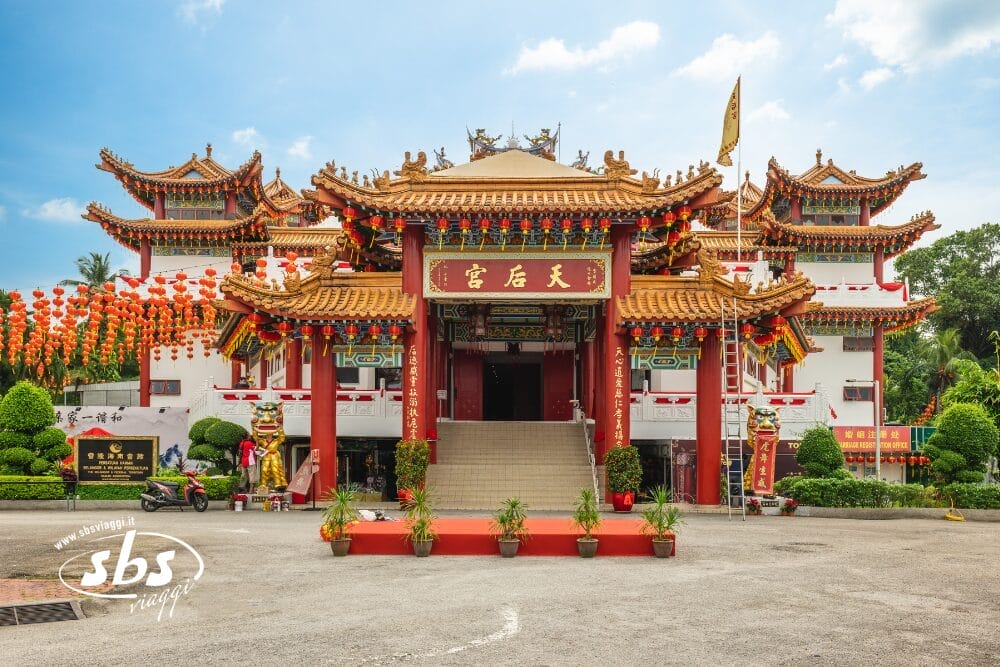 Un tempio cinese tradizionale a Singapore con decorazioni ornamentali rosse e oro, lanterne appese sul lato sinistro e scale che conducono all'ingresso principale sotto un cielo blu. Il tempio è circondato dal verde e irradia un'atmosfera pacifica.