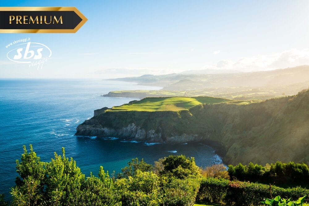 Un paesaggio costiero con scogliere verdi e dolci colline accanto a un mare azzurro e calmo sotto un cielo limpido, visto dall'alto. Il testo "PREMIUM", la filigrana "sbs" e Bozza automatica appaiono nell'immagine.