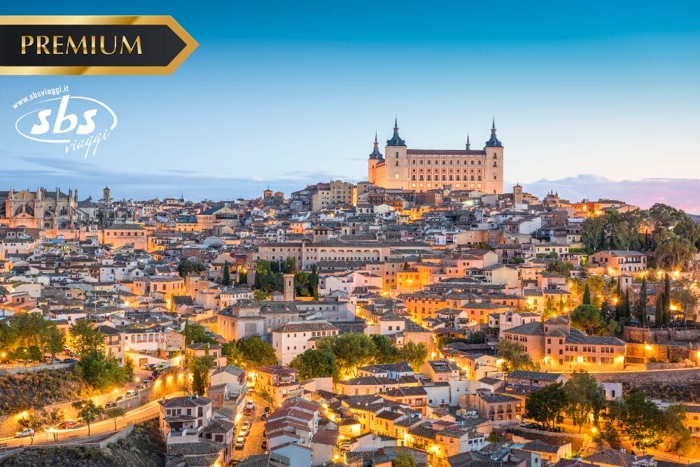 Una vista panoramica di Toledo, in Spagna, al tramonto, con le luci calde che illuminano il paesaggio storico e la grande fortezza dell'Alcázar che domina la città, a breve distanza da Madrid. I loghi "PREMIUM" e "sbs viaggi" appaiono nell'immagine.