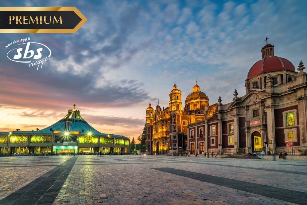 Un'ampia piazza al tramonto con la Basilica di Nostra Signora di Guadalupe illuminata e l'antica basilica di Città del Messico. Nell'angolo in alto a sinistra compaiono l'etichetta "PREMIUM", Bozza automatica e il logo di SBS Viaggi.