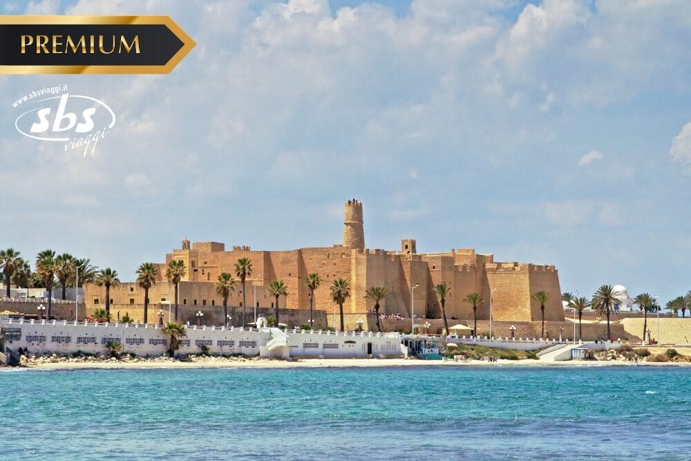 Una grande fortezza storica con alte mura e una torre centrale si erge in riva al mare, costeggiata da palme. Il cielo è azzurro con nuvole. I loghi "PREMIUM", "Bozza automatica" e "sbs viaggi" appaiono nell'angolo superiore sinistro dell'immagine.