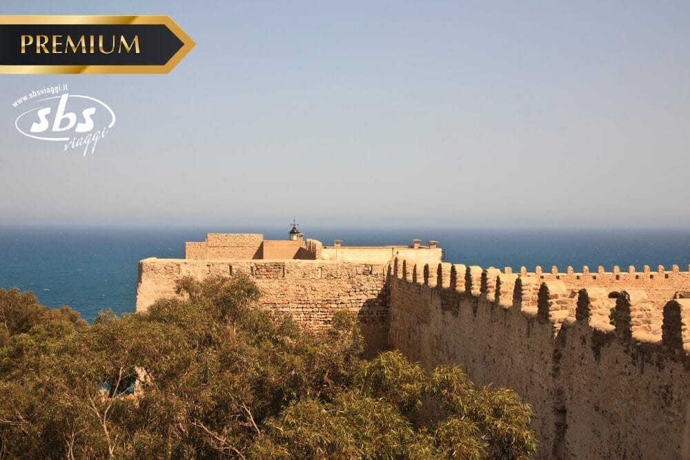 Una fortezza in pietra con mura merlate si affaccia sul mare blu, incorniciata da alberi in primo piano: una scena perfetta del Gran Tour Tunisia. I loghi "PREMIUM" e "sbs viaggi" sono sovrapposti negli angoli superiori dell'immagine.