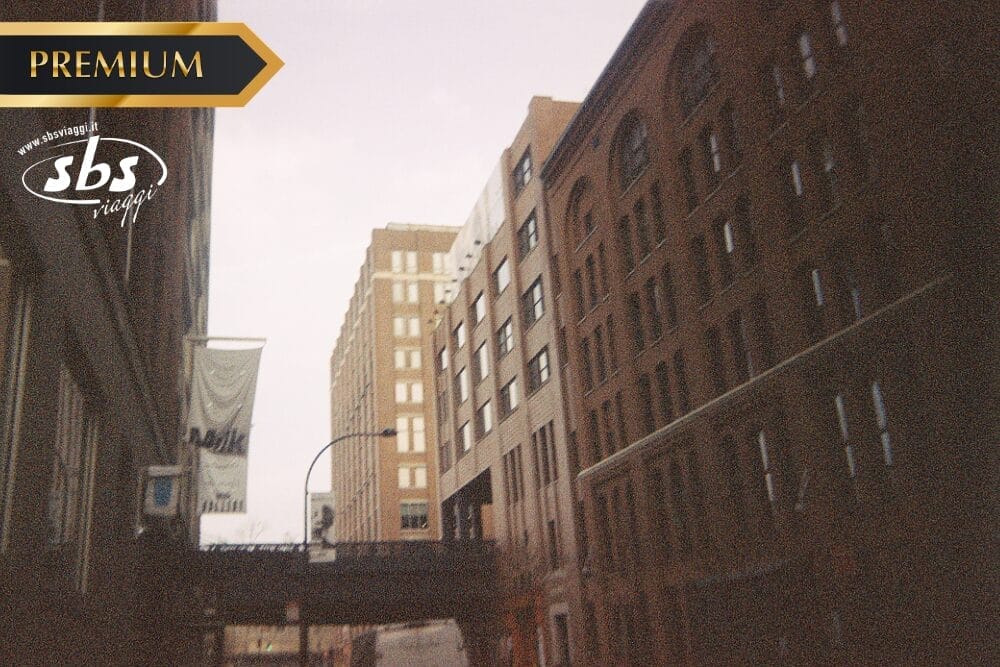 Una scena di strada cittadina con alti edifici in mattoni e pietra, un ponte pedonale e striscioni appesi ai pali della luce. L'immagine, contrassegnata come "PREMIUM", presenta una tonalità calda e vintage e una sottile "Bozza automatica" vicino a "sbs.viaggi".