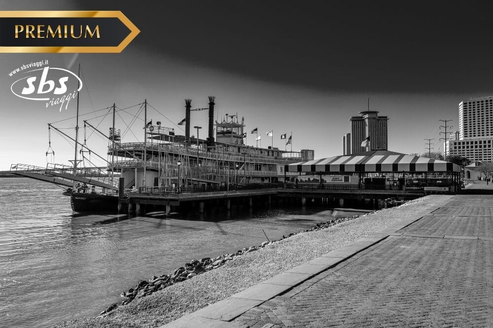 Foto in bianco e nero di un battello fluviale attraccato in riva al mare, vicino a un edificio con tetto a strisce e a un paesaggio urbano. I loghi "PREMIUM", "sbs viaggi" e Bozza automatica sono visualizzati nell'angolo in alto a sinistra.