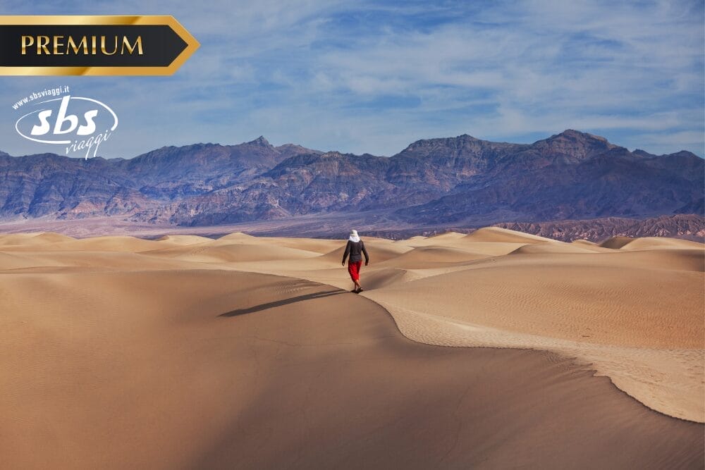 Una persona cammina da sola su dune di sabbia dorata sotto un cielo azzurro, con montagne aspre sullo sfondo. L'immagine presenta le diciture "PREMIUM", "sbs viaggi" e una sottile filigrana Bozza automatica per l'autenticità.