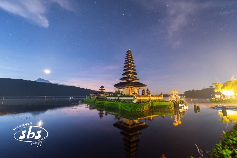 Un tempio tradizionale balinese sorge su un isolotto circondato da acque calme in un cielo crepuscolare vicino a Ubud, con montagne e luna sullo sfondo. Il logo di SBS Viaggi appare nell'angolo in basso a sinistra.