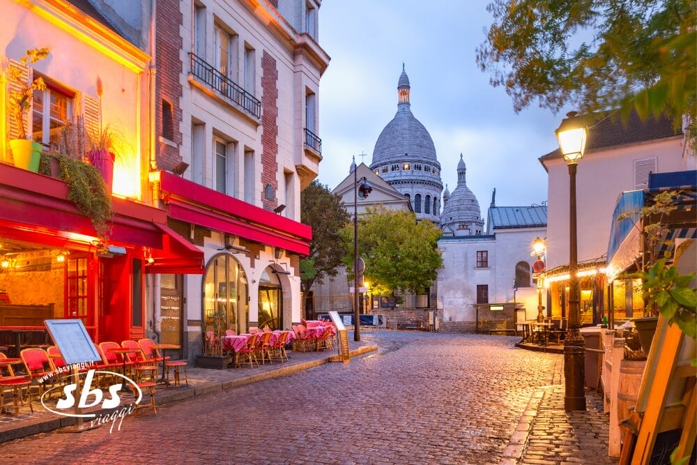 Una tranquilla strada acciottolata di Montmartre, Parigi, con caffè e tavolini colorati all'esterno. Sullo sfondo le cupole bianche della Basilica del Sacro Cuore, in un cielo nuvoloso dell'Île-de-France.
