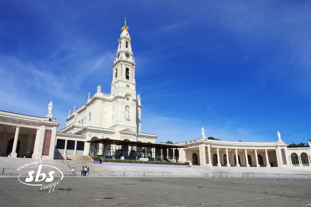 L'immagine mostra il Santuario di Fátima a Portogallo, con la sua basilica centrale alta e bianca con il campanile in un cielo azzurro e limpido, e una grande piazza aperta, una delle Meraviglie del Portogallo.