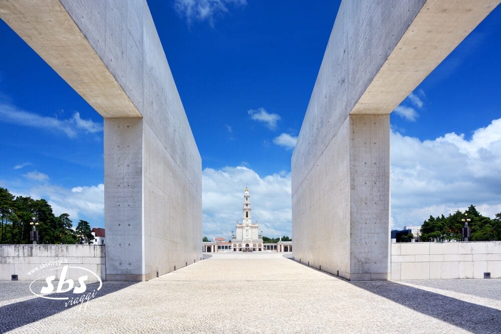 Due grandi e moderni muri di cemento incorniciano un'ampia piazza che conduce al Santuario di Nostra Signora di Fátima, un punto culminante di qualsiasi tour di Portogallo, con la sua chiesa bianca e l'alto campanile sotto un cielo azzurro brillante con nuvole sparse.