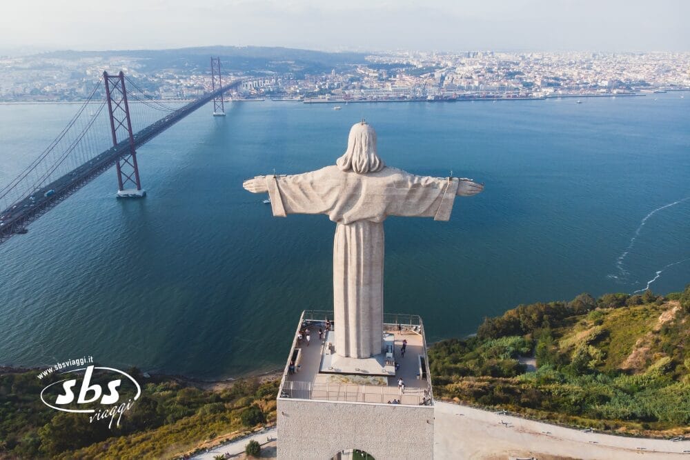 Veduta aerea della statua di Cristo Re ad Almada, Sud del Portogallo, con vista sul fiume Tago e sul Ponte 25 de Abril, con Lisbona visibile sullo sfondo e il logo di SBS Viaggi in basso a sinistra.