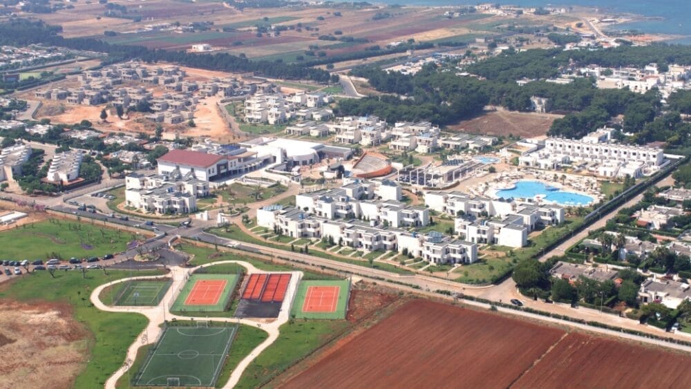 Vista aerea di un grande complesso turistico con edifici bianchi, piscina, campi da tennis e da calcio, circondato dal verde e da campi agricoli: questa Bozza automatica offre una vista sulla costa e altri edifici in lontananza.