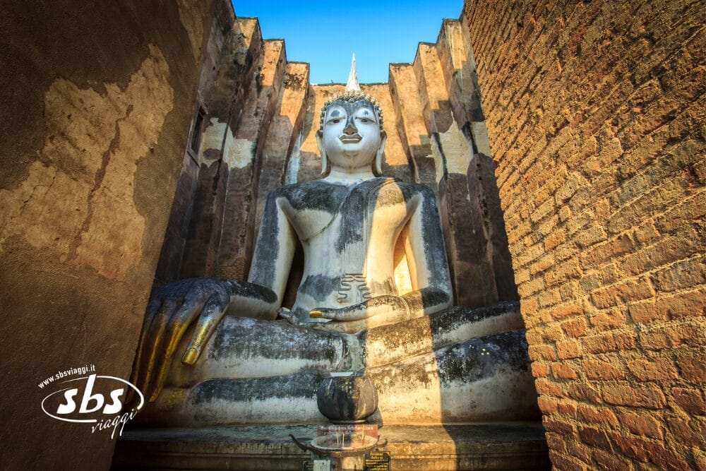 Una grande statua di Buddha seduta, con il volto e le mani bianche, è incorniciata da antiche mura di mattoni sotto un cielo azzurro: una vista iconica di ogni viaggio in Thailandia. L'espressione serena e le dita dorate spiccano, con il logo di SBS Viaggi nell'angolo in basso a sinistra.