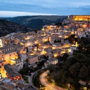 Una città collinare al tramonto, con strade tortuose ed edifici in pietra illuminati da caldi lampioni. Il paesaggio è caratterizzato da dolci colline e da una grande struttura illuminata in cima, come una bozza luminosa automatica contro il cielo notturno.