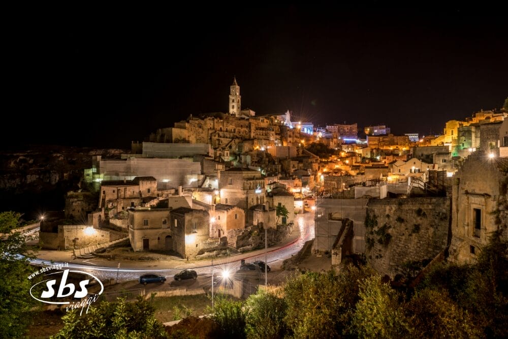Veduta notturna di Matera, Italia, con edifici storici in pietra illuminati e un campanile su una collina. Strade tortuose e auto parcheggiate riempiono il primo piano. Il logo SBS Travel e "Bozza automatica" appaiono nell'angolo in basso a sinistra.