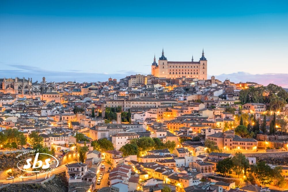 Una vista panoramica di Toledo, in Spagna, al tramonto, con edifici storici e case illuminate a giorno. L'imponente Alcázar di Castiglia si staglia in cima alla collina, in un cielo azzurro e limpido: una meta perfetta per il Minitour di Pasqua.