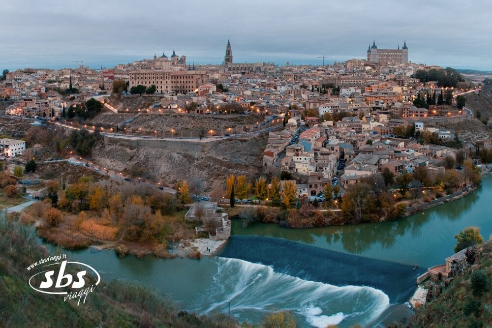 Una vista panoramica di Toledo, in Spagna, che mostra edifici storici e una cattedrale su una collina, con il fiume Tago e una piccola cascata in primo piano in una giornata nuvolosa, ripresa con Bozza automatica per una chiarezza senza soluzione di continuità.