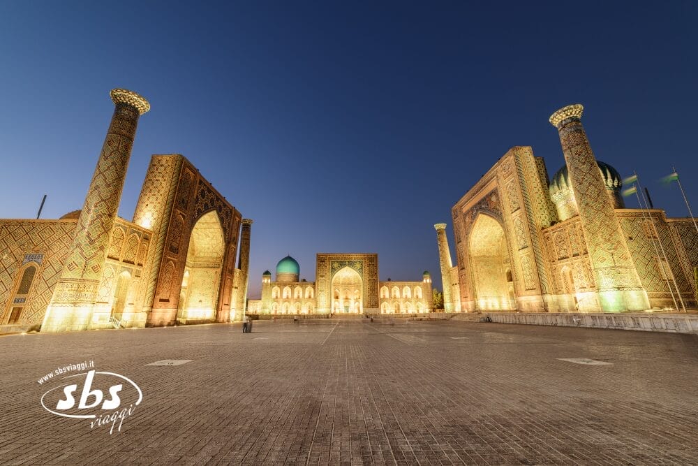 Un'ampia piazza vuota al tramonto, circondata da tre grandiose madrase illuminate con intricati motivi a mosaico e alti minareti nella piazza Registan di Samarcanda, in Uzbekistan: una scena incantevole degna di una Bozza automatica.