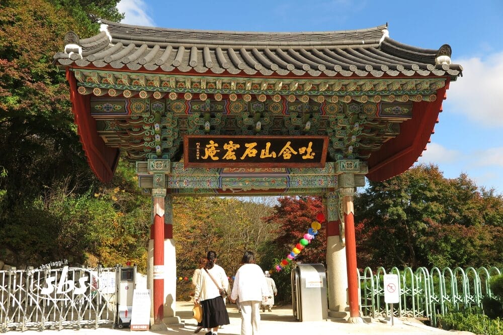 Due persone attraversano un colorato cancello coreano tradizionale in Corea del Sud, ornato da intricati disegni e scritte asiatiche, circondato da fogliame autunnale e da un cielo azzurro: una tappa perfetta di un Tour in libertà.