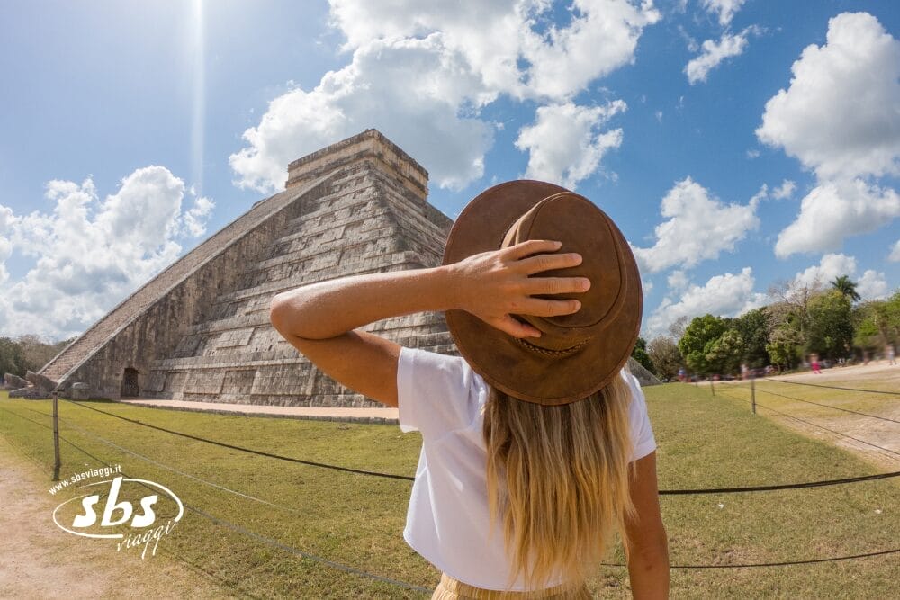 Una persona con lunghi capelli biondi che indossa un cappello marrone e una camicia bianca si trova di fronte all'antica piramide di Chichen Itza nello Yucatán, in Messico, sotto un cielo parzialmente nuvoloso, con le spalle alla macchina fotografica e la mano appoggiata sul cappello.
