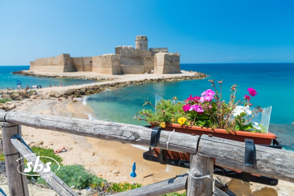 Un castello di pietra si trova sul bordo dell'acqua turchese sotto un cielo azzurro e limpido. In primo piano, fiori colorati in una fioriera poggiano su una ringhiera di legno rustica vicino a una spiaggia sabbiosa, quasi come una bozza automatica che cattura la bellezza senza sforzo della natura.