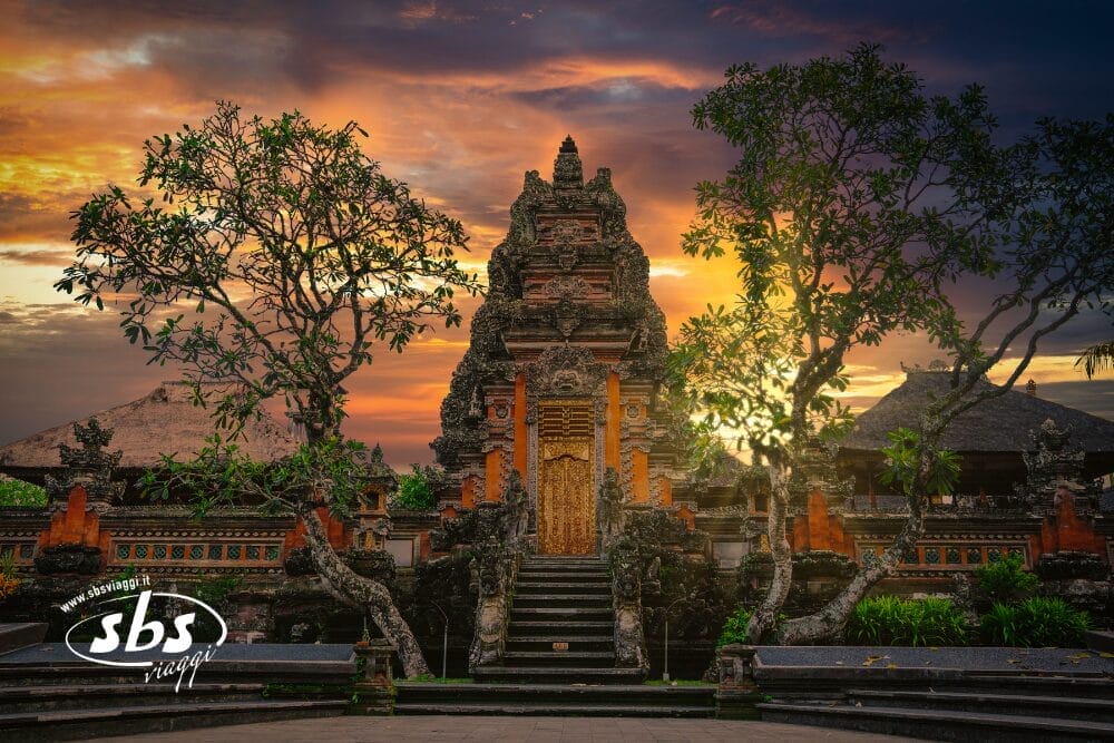 Un tradizionale tempio balinese con intricate sculture in pietra, circondato da alberi, è illuminato da un drammatico cielo arancione e viola al tramonto. L'ingresso ornato evoca il fascino storico di Bali, a breve distanza da Singapore.