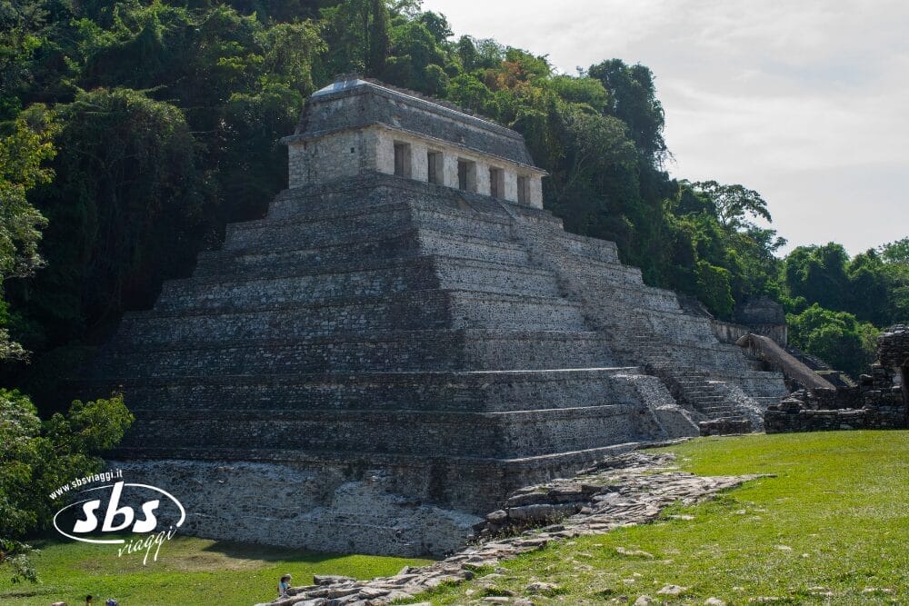 Una grande e antica piramide di pietra nello Yucatán si erge al di sopra di fitti alberi verdi sotto un cielo parzialmente nuvoloso, con la luce del sole che proietta ombre sui gradini e su un'area erbosa in primo piano.