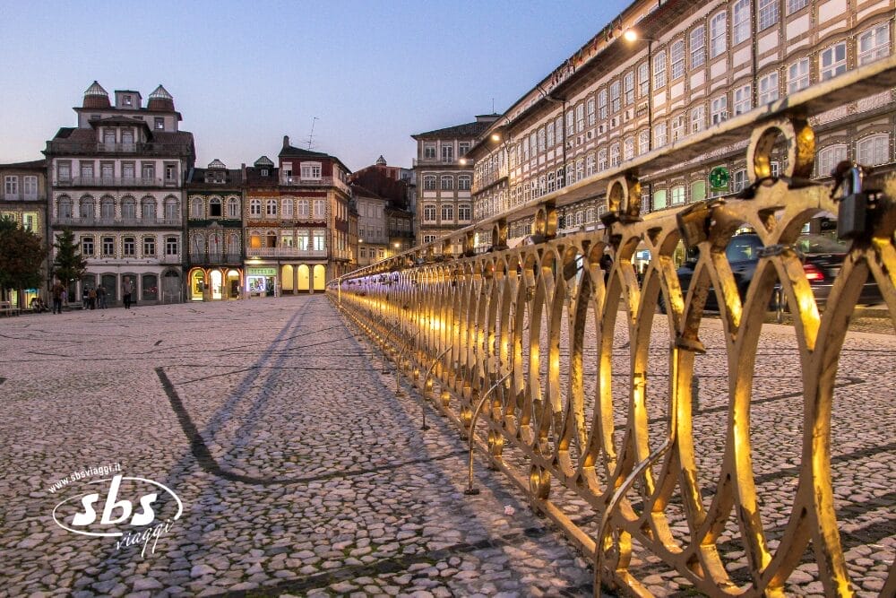 Una piazza acciottolata al tramonto con ringhiere dorate, edifici storici con finestre illuminate e auto che passano sullo sfondo. Bozza automatica appare sottilmente mentre il logo "sbs" è visualizzato nell'angolo in basso a sinistra.