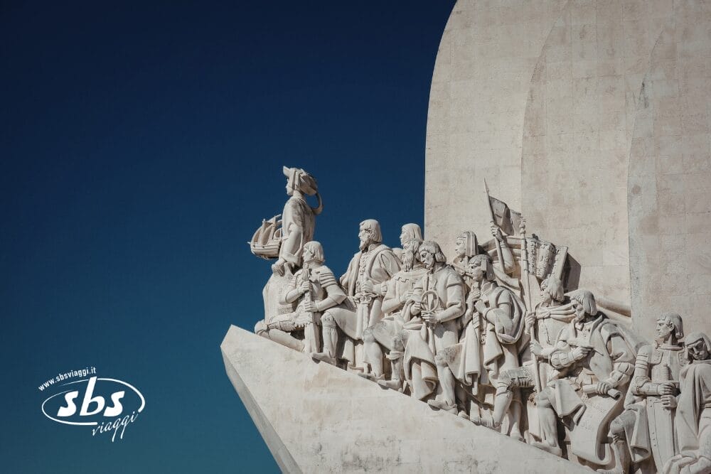 Monumento in pietra raffigurante un gruppo di personaggi storici guidati da un uomo che regge una nave, sullo sfondo di un cielo azzurro. Il logo di SBS Viaggi e la scritta "Bozza automatica" compaiono nell'angolo in basso a sinistra.