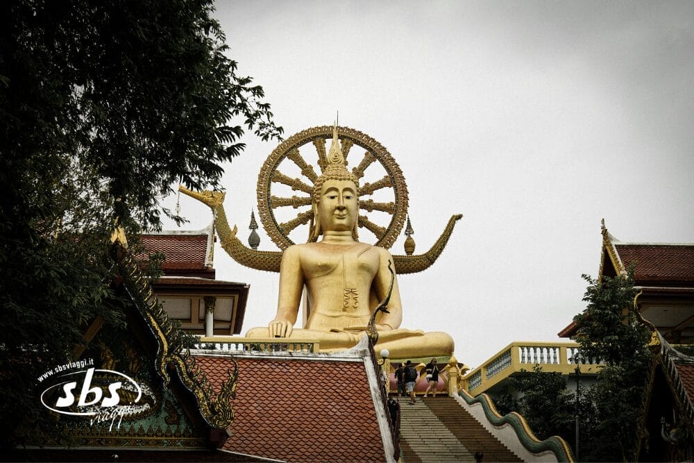 Una grande statua dorata di Buddha si trova in cima a una scalinata di Siem Reap, con decorazioni ornamentali e un'aureola circolare dietro la testa, affiancata dai tetti dei templi e dal verde. Sono visibili persone che salgono le scale.