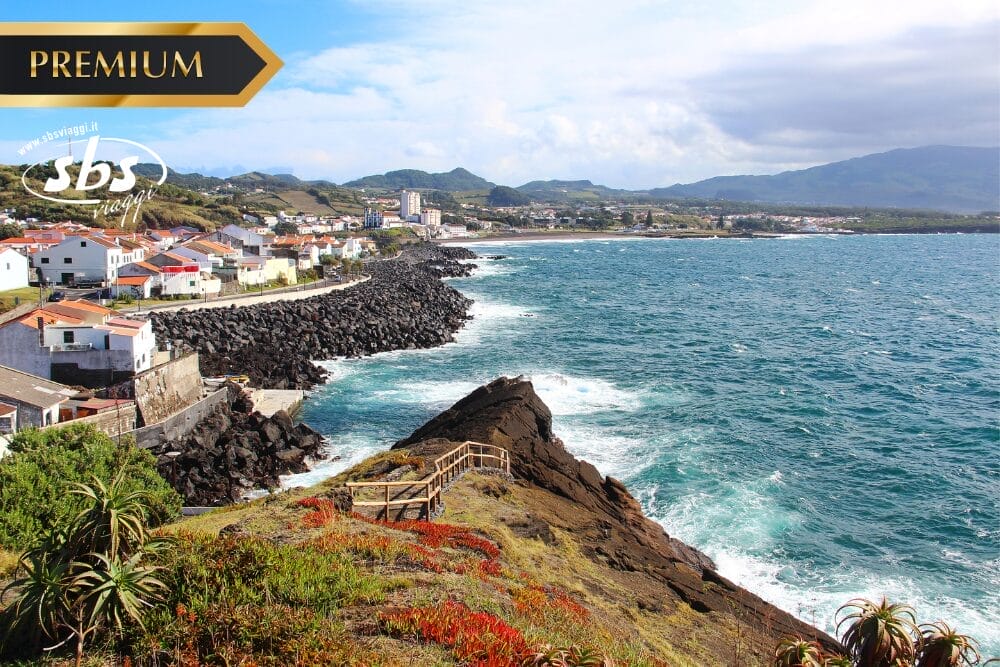 Una città costiera con case bianche e tetti rossi confina con una costa rocciosa e l'oceano blu, con montagne e verde sullo sfondo sotto un cielo parzialmente nuvoloso. Una scala di legno scende dalle rocce. In alto a sinistra compaiono le scritte "PREMIUM" e "Bozza automatica".
