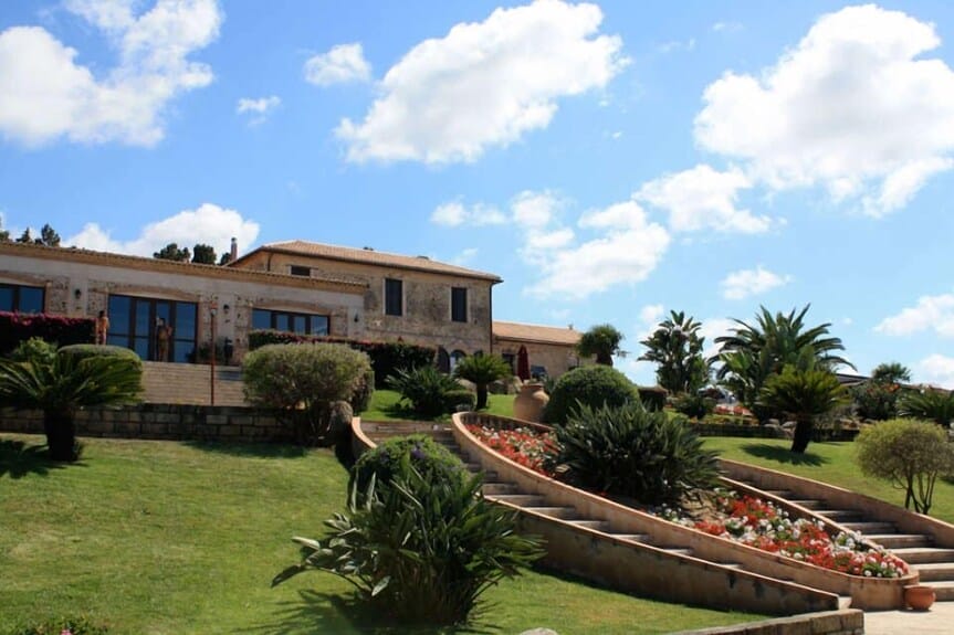 Una grande villa in pietra si trova su una collina erbosa, circondata da giardini, palme e aiuole vivaci sotto un cielo azzurro con nuvole sparse: una vera Bozza automatica di bellezza naturale.