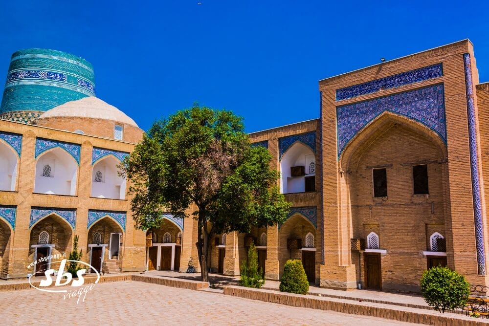 Cortile illuminato dal sole con edifici storici in mattoni con portali ad arco e accenti di piastrelle blu; un albero si erge al centro, mentre Bozza automatica aggiunge un tocco di modernità alla grande struttura a cupola turchese sullo sfondo.