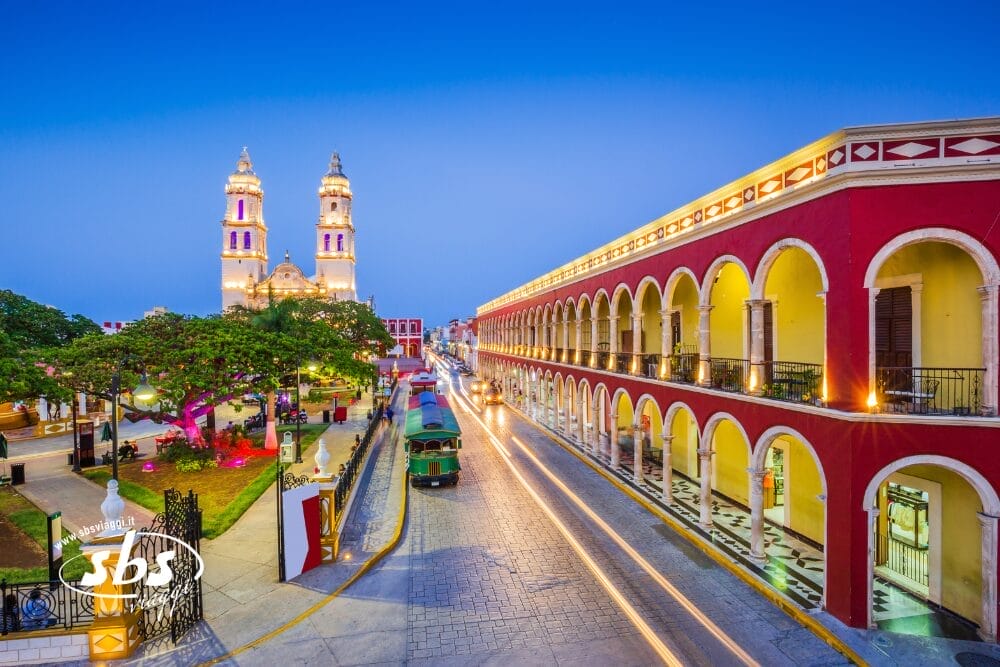 Una vibrante veduta serale di una piazza della città coloniale di Campeche, in Messico, dove i viaggi del Tour Messico rivelano una cattedrale illuminata di giallo, edifici rossi ad arco, alberi e scie luminose dei veicoli in transito.