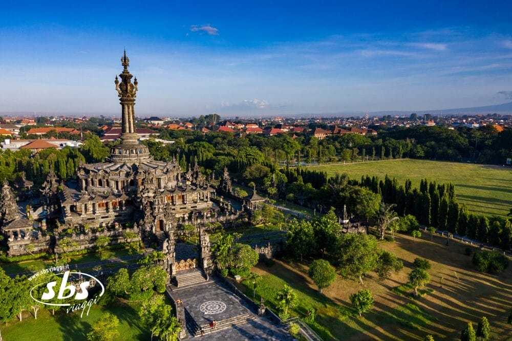 Veduta aerea di un grande monumento con intricati intagli circondato da verde e alberi a Bali, situato in un'area urbana con molti edifici dai tetti rossi sotto un cielo azzurro e limpido.
