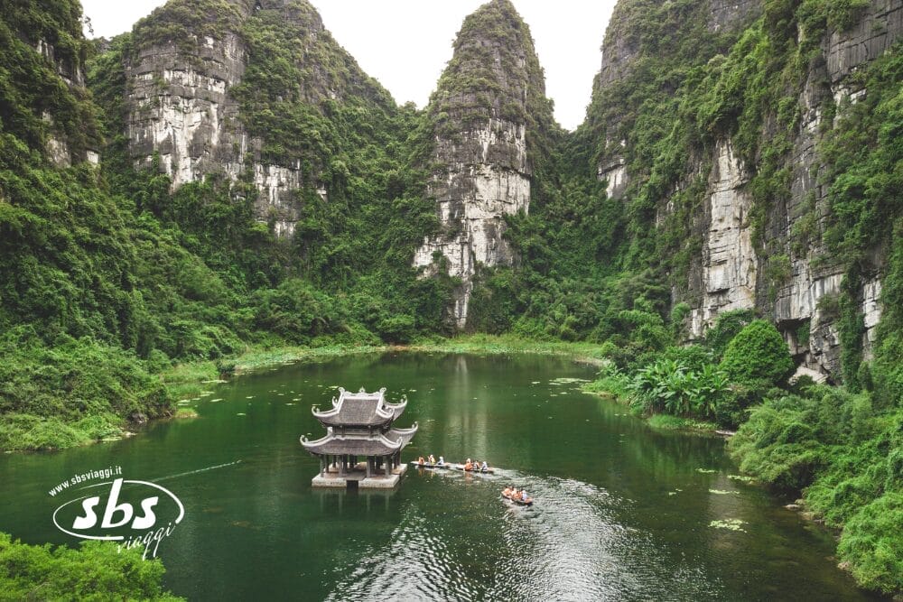 Un padiglione tradizionale in legno si trova su un tranquillo lago verde circondato da ripide e rigogliose scogliere calcaree. Un gruppo di persone in barca a remi - parte dei tour de Le Perle dell'Indocina - si avvicina al padiglione in mezzo a una fitta e spettacolare vegetazione.